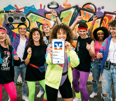 Junge Menschen im 80er Jahre-Look halten ein Handy in die Kamera auf dem die VR Banking App zu sehen ist.