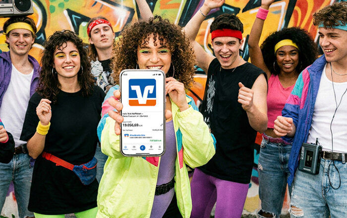 Junge Menschen im 80er Jahre-Look halten ein Handy in die Kamera auf dem die VR Banking App zu sehen ist.